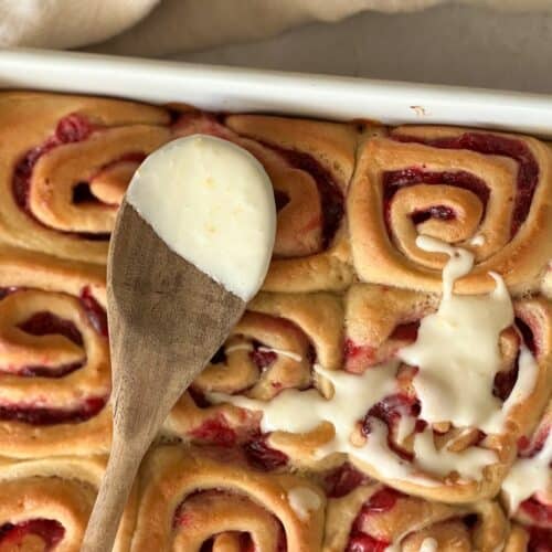 cranberry orange rolls being spread with frosting using a wooden spoon.