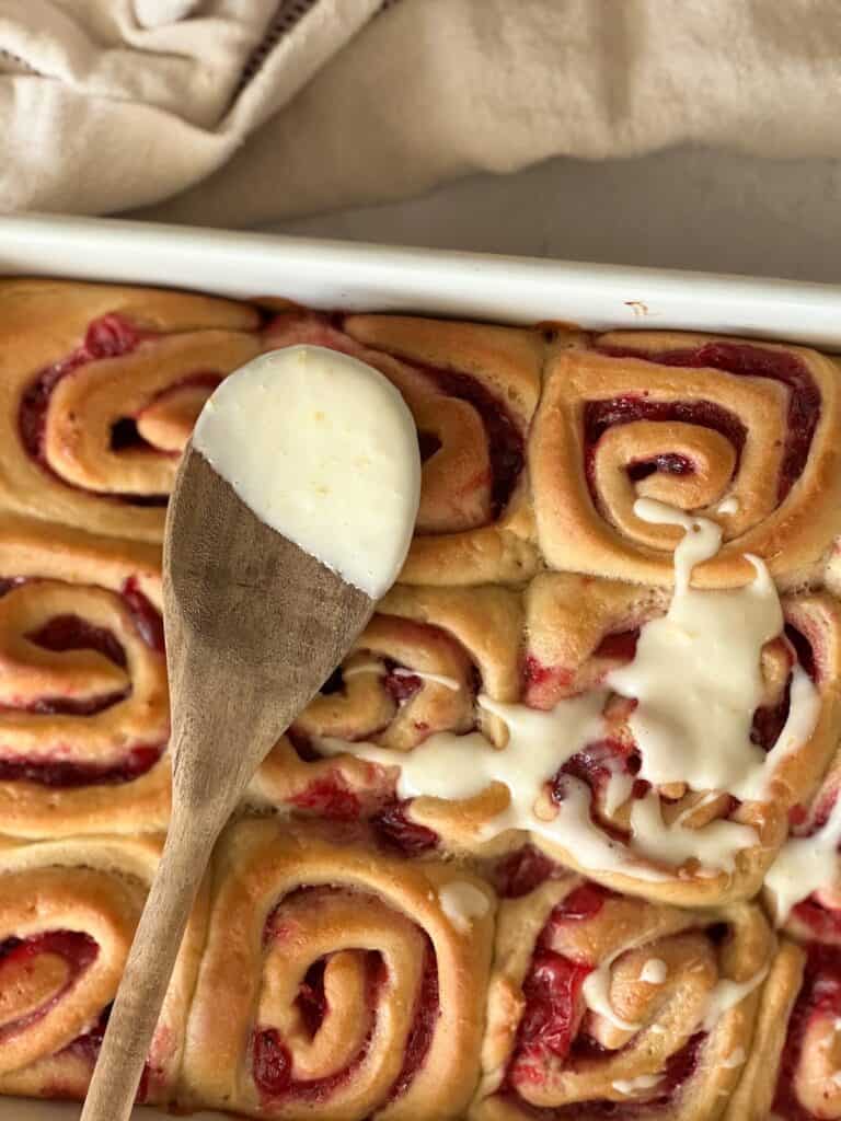 cranberry orange rolls being spread with frosting using a wooden spoon.