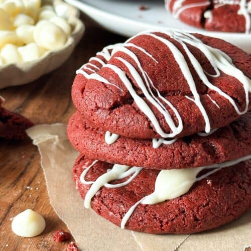 There are three red velvet sourdough cookies stacked on top of each other with a bowl of white chocolate chips and a plate of more cookies in the background.