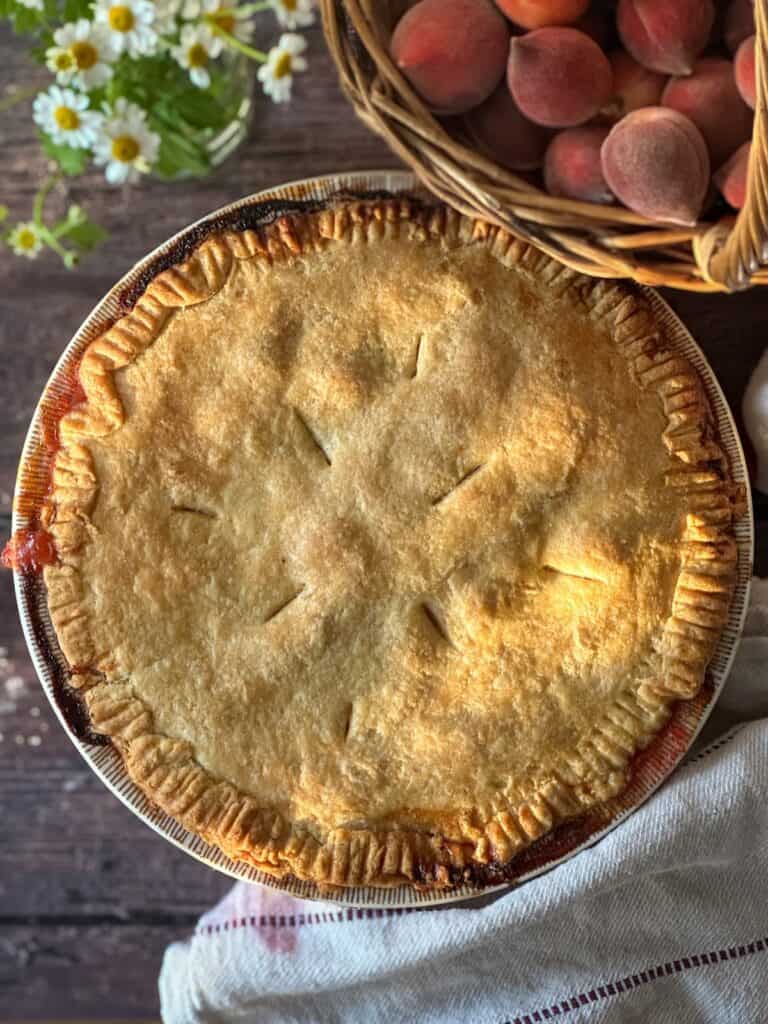 whole sourdough peach pie next to a basket of fresh peaches.