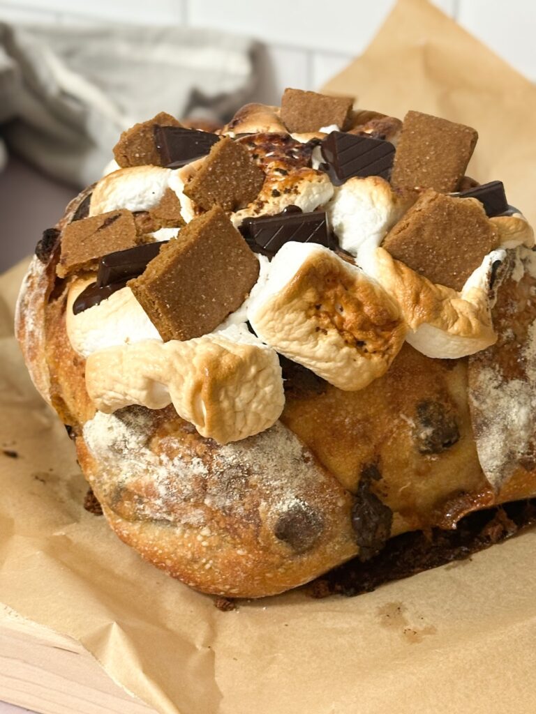 A loaf of s'mores sourdough bread topped with roasted marshmallows, graham crackers and chocolate pieces.