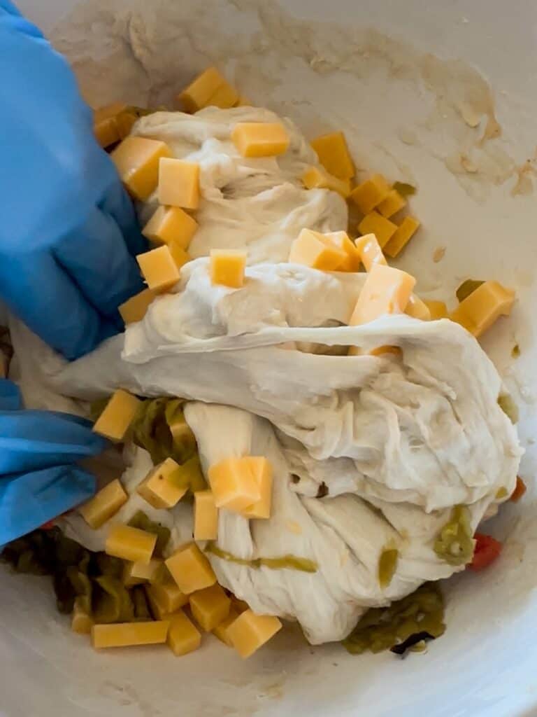 A set of hands wearing blue gloves performing a set of stretch and folds to mix in green chile and cheddar into sourdough dough. 